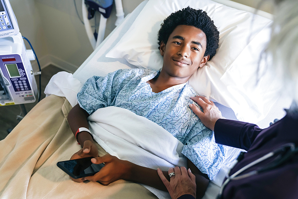 Doctor comforting boy laying in hospital bed holding cell phone