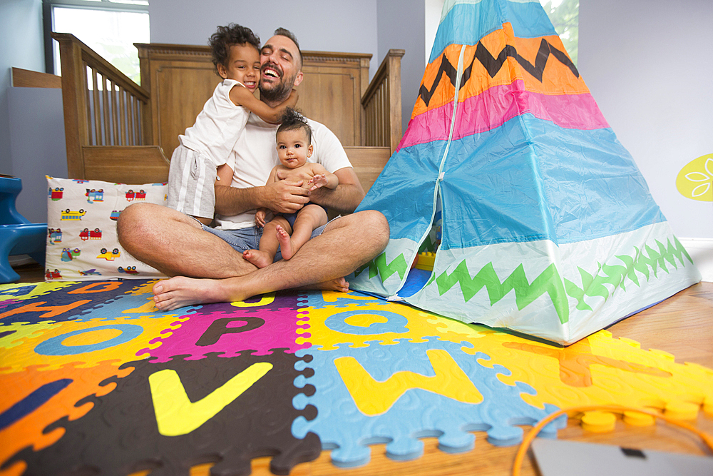 Father sitting on playroom floor holding son and daughter