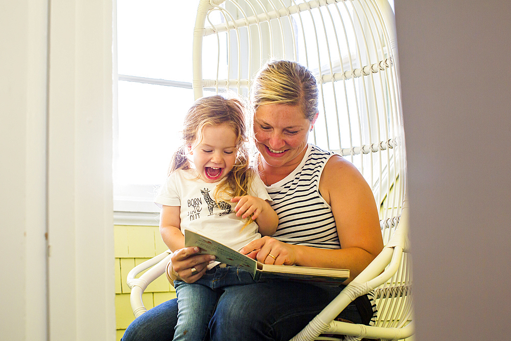 Caucasian mother reading book to excited daughter sitting on lap