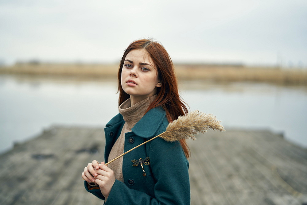 Serious Caucasian woman standing on dock holding stalk of grass