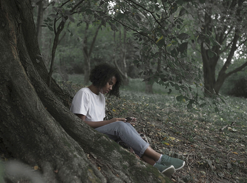 Woman sitting on tree root listening to cell phone with earbuds