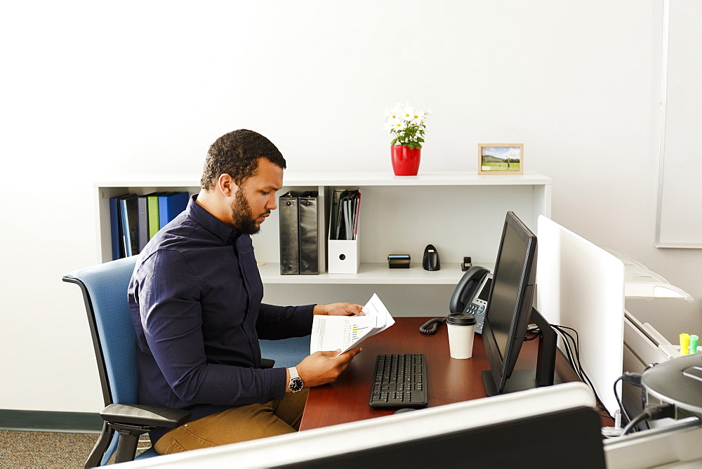 Mixed Race man reading paperwork near computer in office