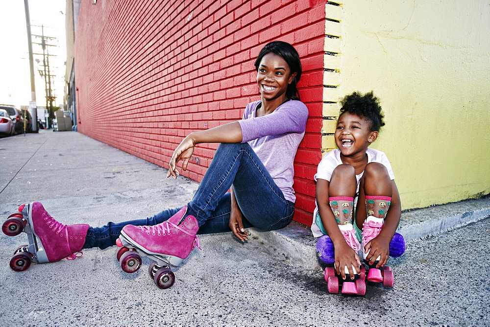 Black mother and daughter wearing roller skates sitting on sidewalk