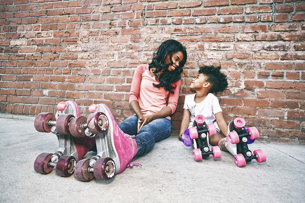 Black mother and daughter wearing roller skates sitting on sidewalk