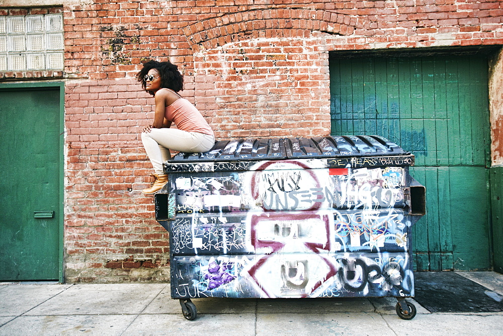 Black woman sitting on dumpster covered with graffiti