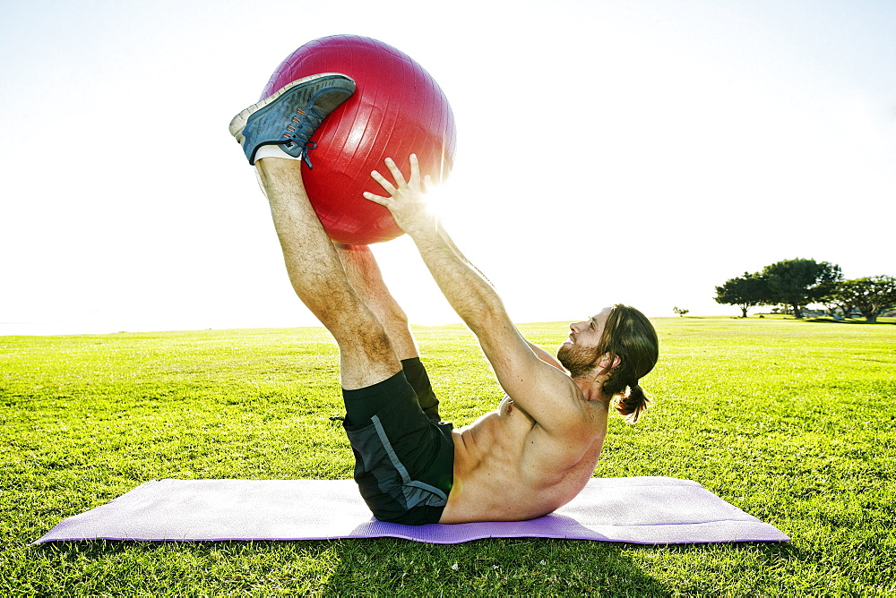 Caucasian man lifting fitness ball with legs in sunny field