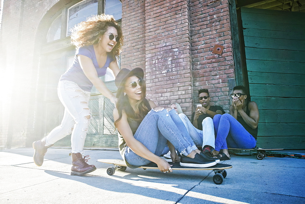 Woman pushing friend sitting on skateboard in city