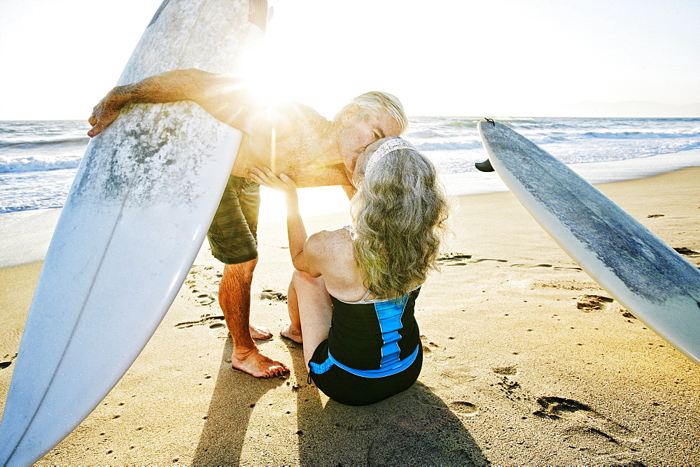 Older Caucasian couple on beach with surfboards kissing