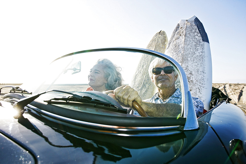 Older Caucasian couple in convertible car with surfboards on beach