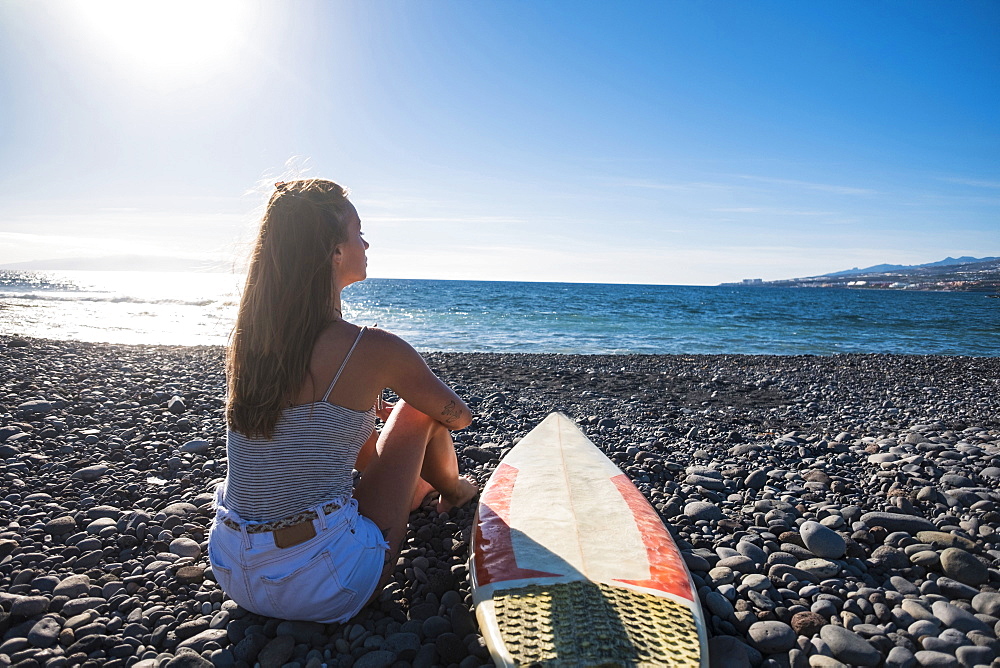 Caucasian woman sitting on beach near surfboard