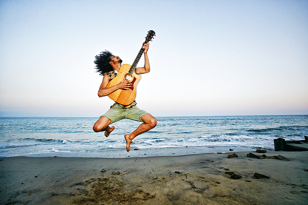 Mixed Race man playing guitar and jumping at beach