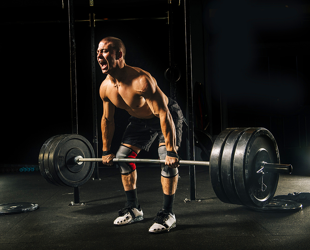 Man screaming while lifting heavy barbell in gymnasium