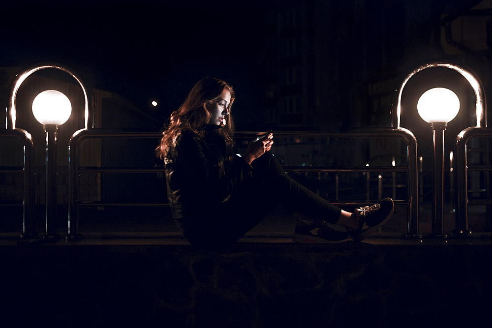 Caucasian teenage girl sitting on bench at night texting on cell phone