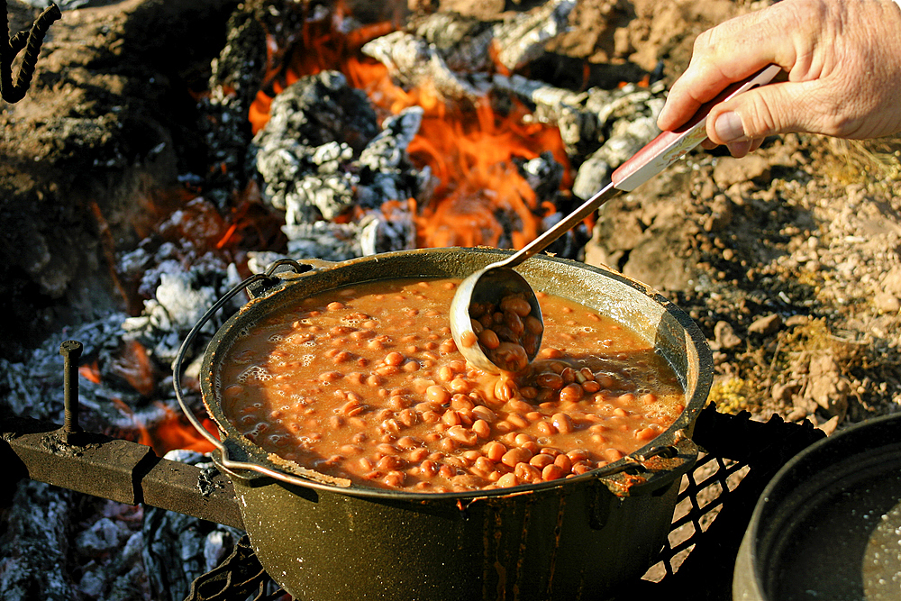 Hand of man stirring beans with ladle on campfire