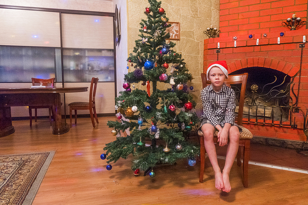 Caucasian boy sitting on chair wearing Santa hat near Christmas tree