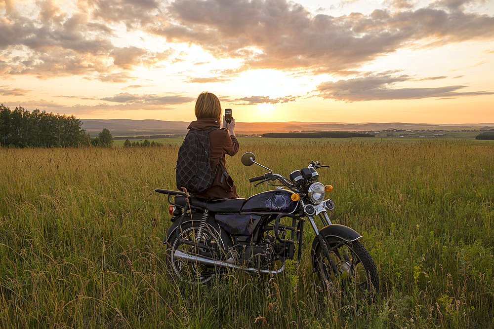 Caucasian woman with motorcycle in field photographing sunset