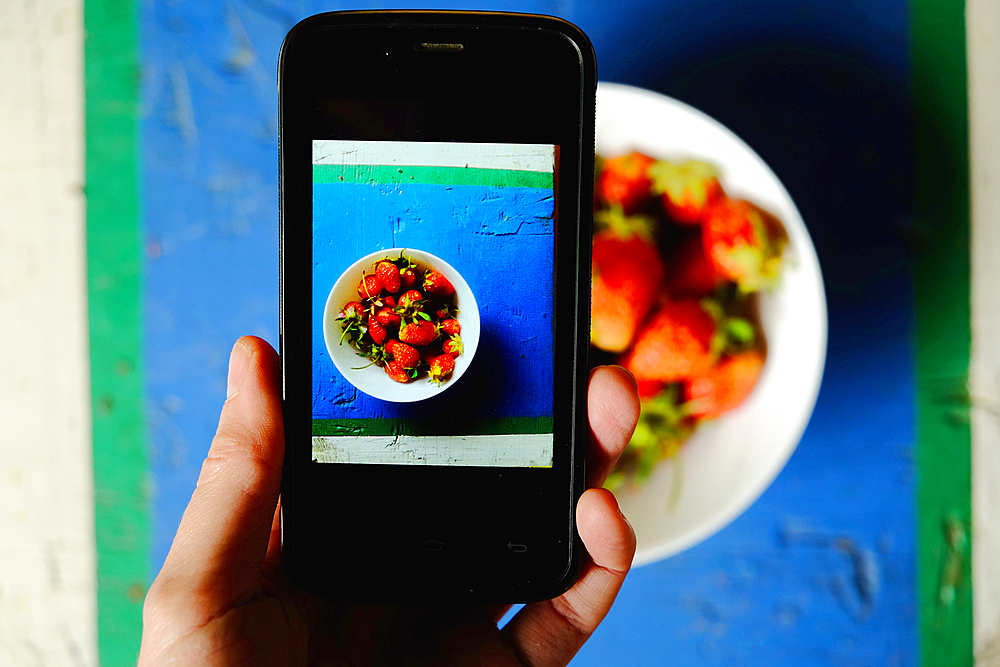 Hand of Caucasian woman photographing bowl of strawberries