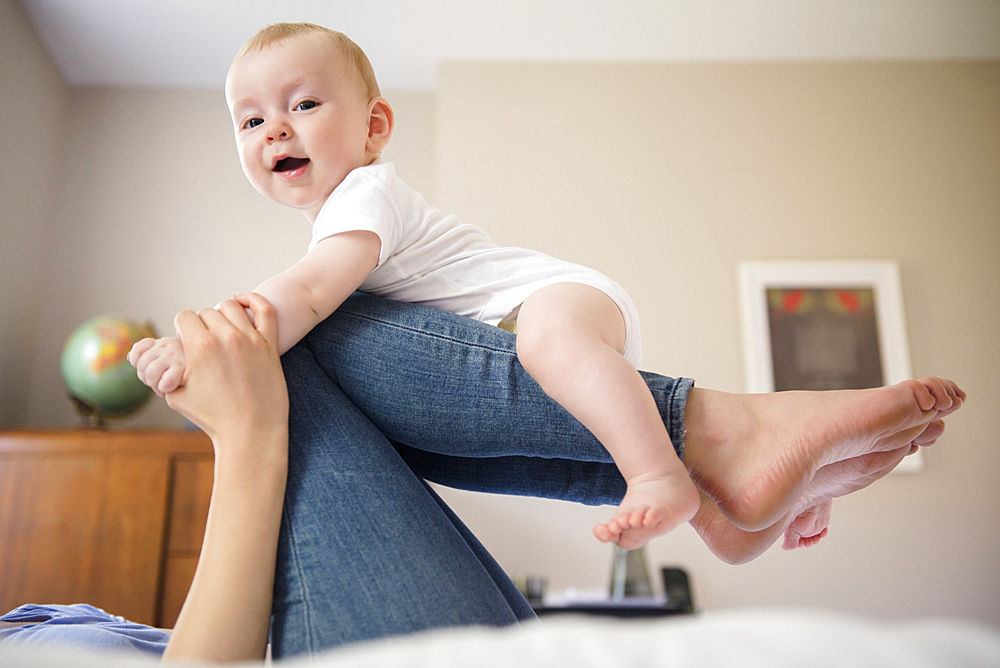 Caucasian mother laying on bed balancing baby son on legs