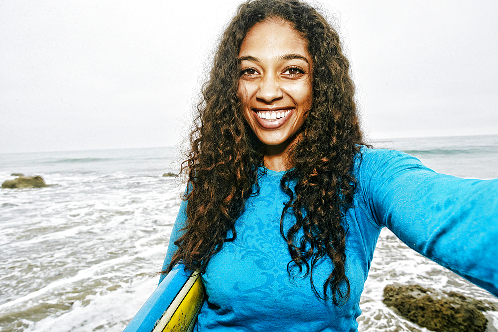 Smiling Mixed Race woman holding surfboard posing for selfie at beach