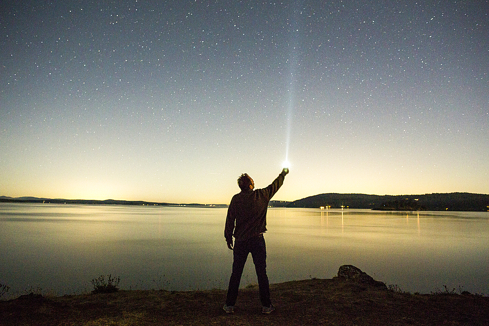 Caucasian man pointing flashlight at night sky near water