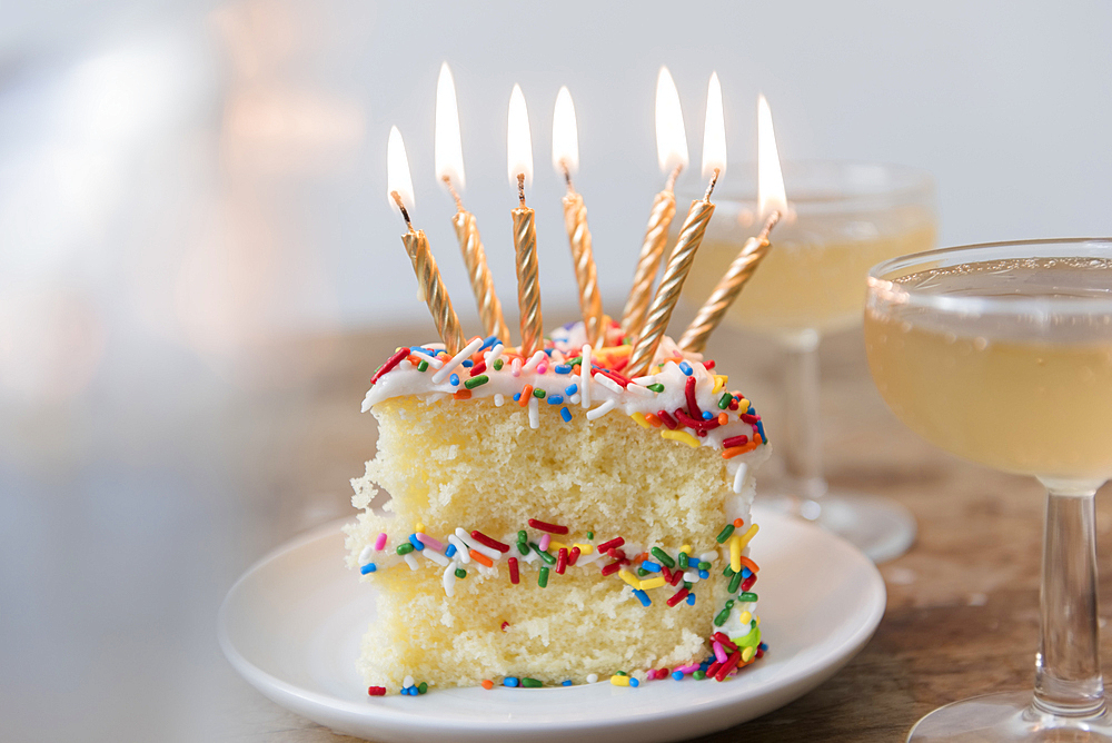 Candles burning on slice of cake with sprinkles near champagne