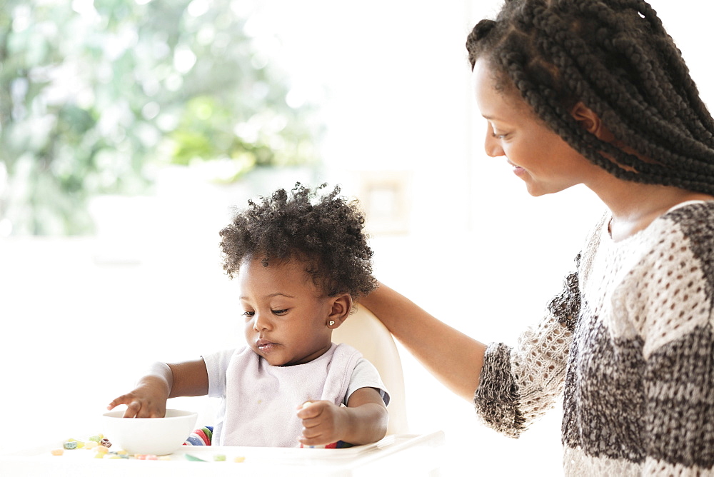 Black woman watching baby daughter eat cereal in high chair