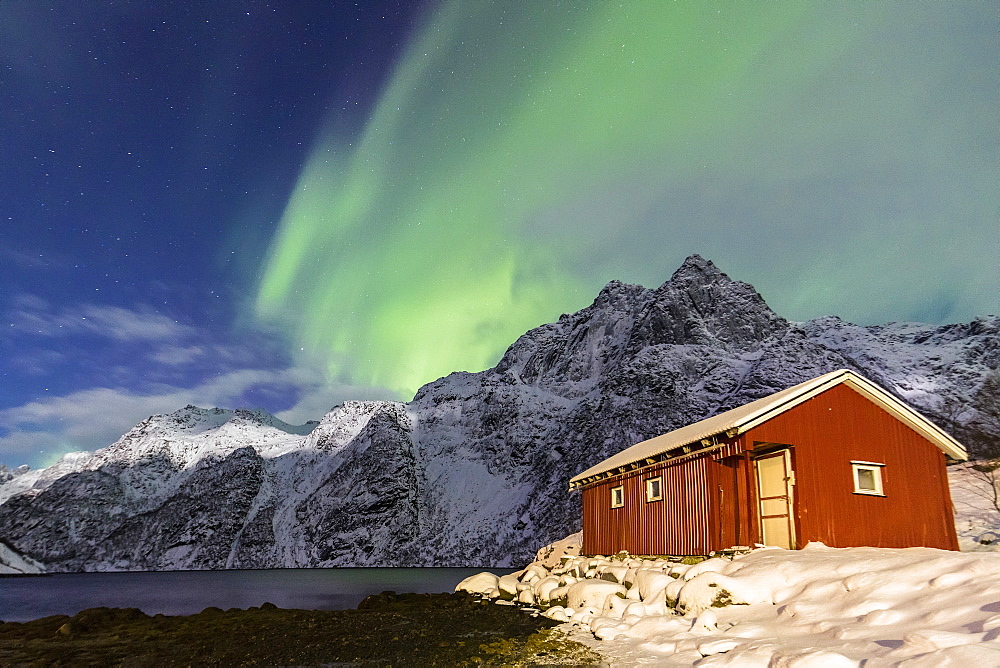 Northern Lights (aurora borealis) illuminate snowy peaks and the wooden cabin on a starry night at Budalen, Svolvaer, Lofoten Islands, Arctic, Norway, Scandinavia, Europe