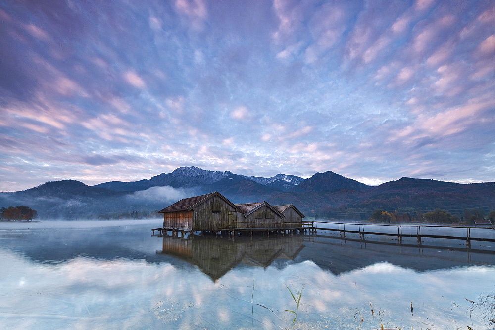 Pink clouds at sunset and wooden huts are reflected in the clear water of Kochelsee, Schlehdorf, Bavaria, Germany, Europe