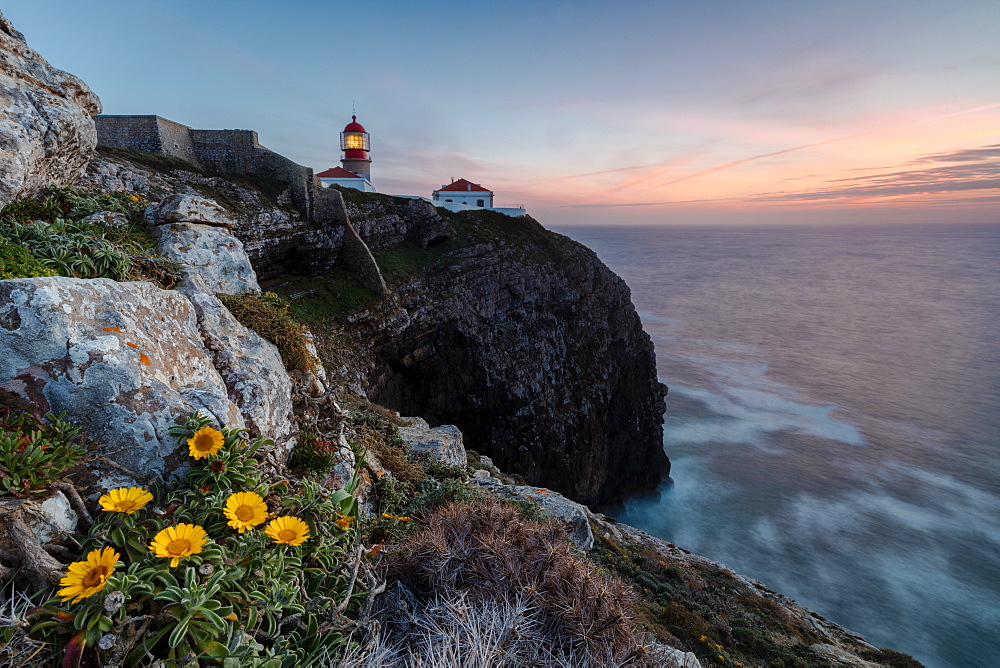 Pink sky at sunset and yellow flowers frame the lighthouse, Cabo De Sao Vicente, Sagres, Algarve, Portugal, Europe