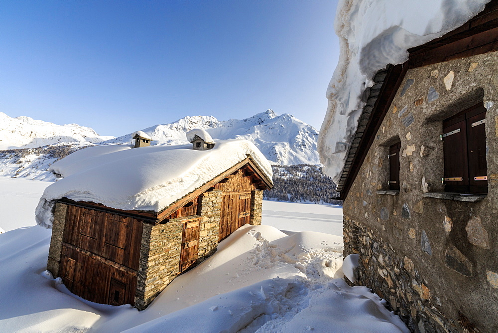 The sun, covered with thin clouds, illuminating a typical hut covered with snow at the Maloja Pass, Graubunden, Swiss Alps, Switzerland, Europe