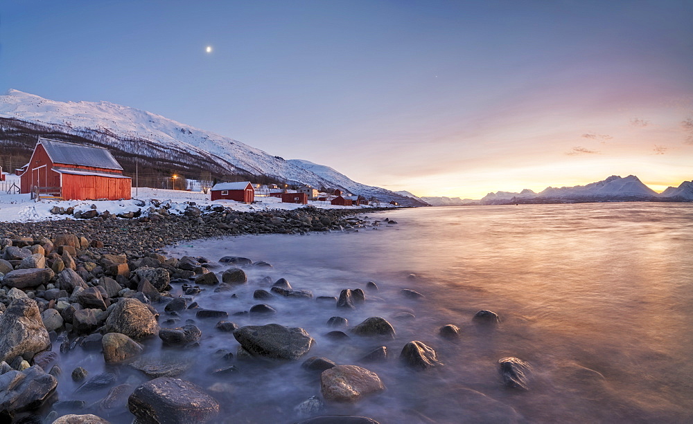 Panorama of typical wood huts called Rorbu framed by fiery sky at sunset and frozen sea, Djupvik, Lyngen Alps, Troms, Norway, Scandinavia, Europe