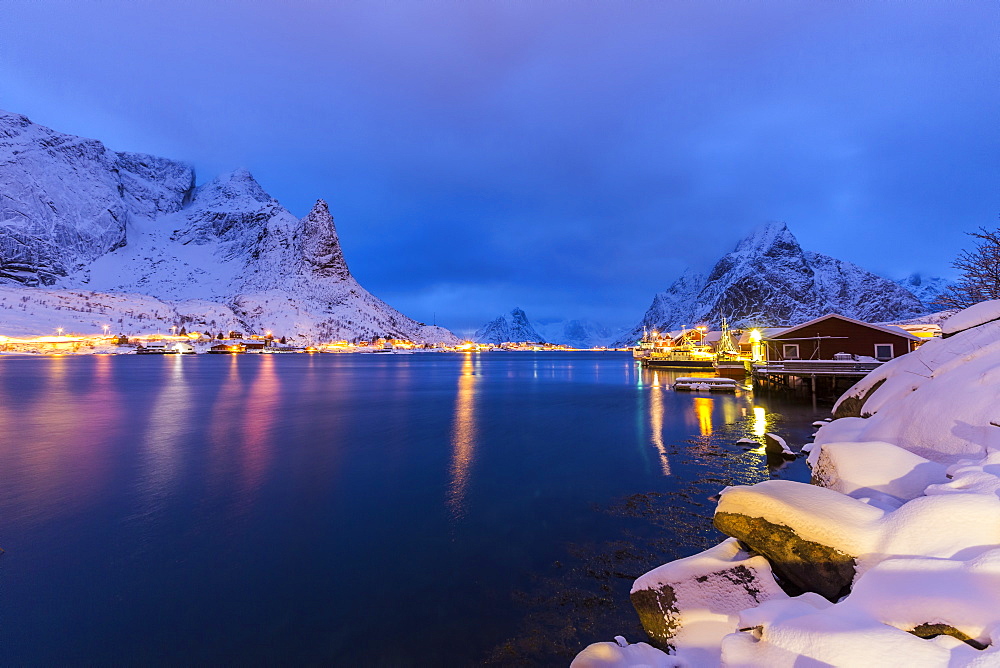 Blue hour in the small bay of Reine, where the lights of the houses reflect in the calm water surrounding the Lofoten Islands, Arctic, Norway, Scandinavia, Europe