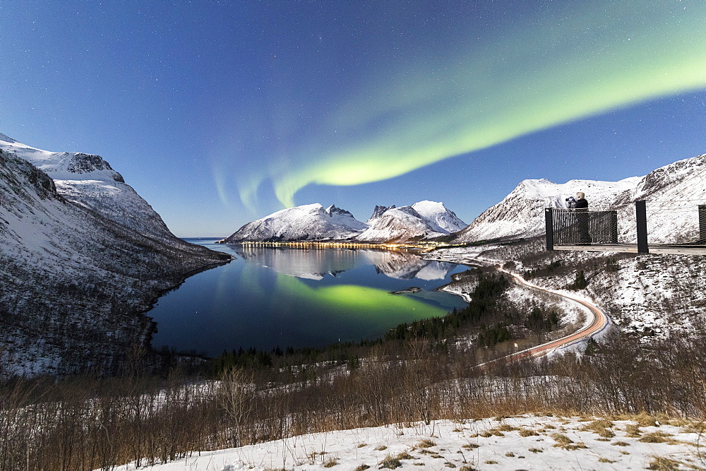 Photographer on platform admires the Northern lights (aurora borealis) and stars reflected in the cold sea, Bergsbotn, Senja, Troms, Norway, Scandinavia, Europe