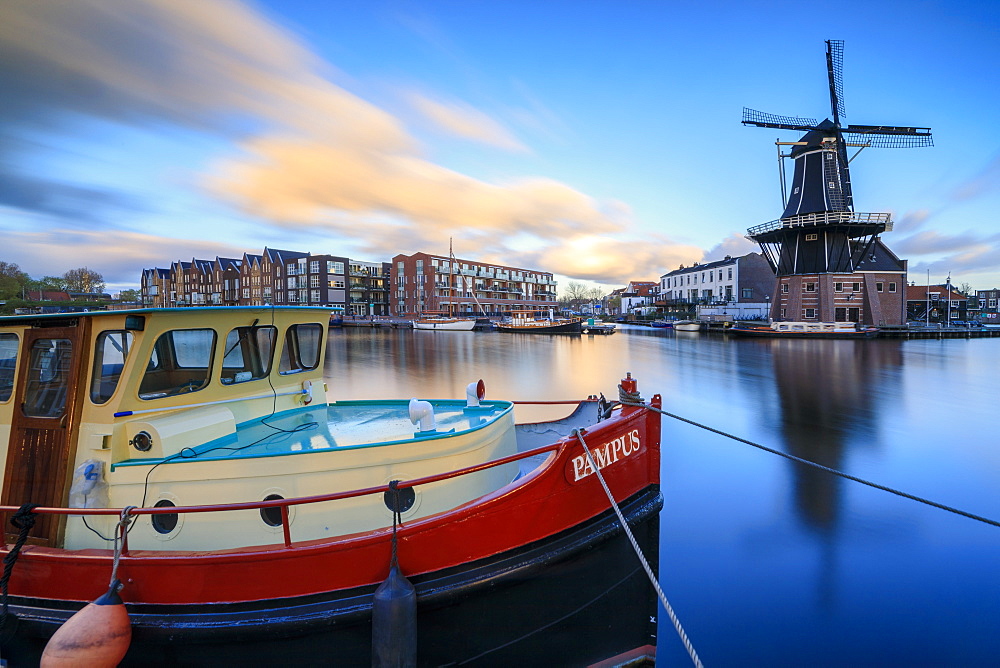 The fishing boat frames the Windmill De Adriaan reflected in the River Spaarne at dusk, Haarlem, North Holland, The Netherlands, Europe