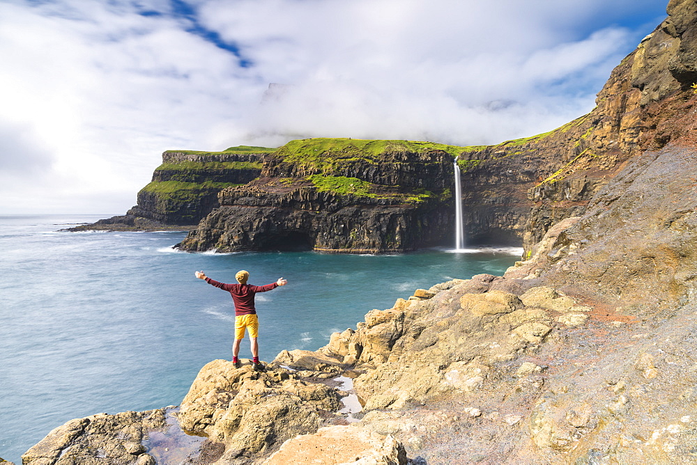 Man on cliffs with open arms admiring Gasadalur waterfall, Vagar island, Faroe Islands, Denmark, Europe