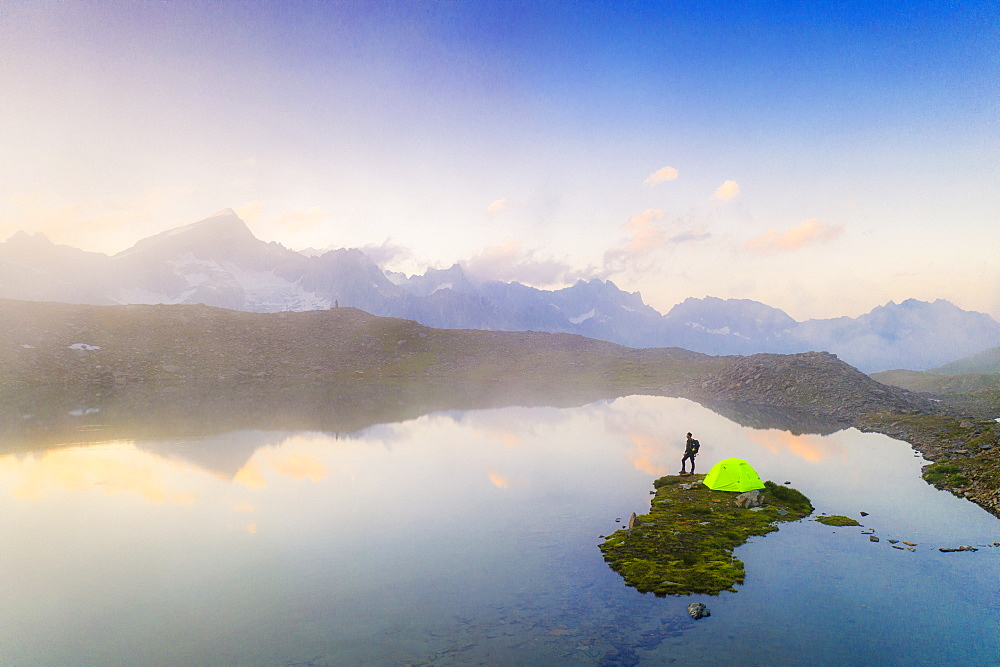 Hiker man with tent admiring a misty sunrise over mountains from Obere Schwarziseeli lake, Furka Pass, Canton Uri, Switzerland, Europe