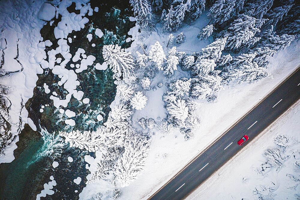 Car traveling on the snowy mountain road on side of frozen river and woods, aerial view, Switzerland, Europe