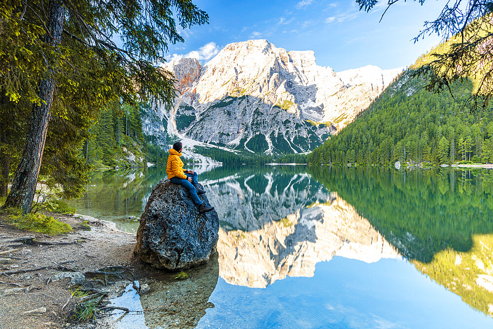 Hiker man enjoying sunrise sitting on rocks on shore of Lake Braies (Pragser Wildsee) at dawn, Dolomites, South Tyrol, Italy, Europe