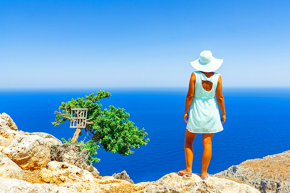 Back view of woman with fashion dress and hat looking at the sea from cliffs, Crete, Greek Islands, Greece, Europe