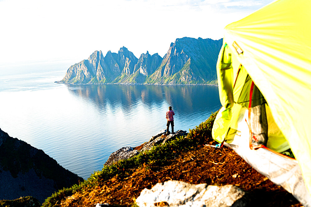 Hiker admiring mountains standing on top of cliff beyond camping tent, Senja island, Troms county, Norway, Scandinavia, Europe