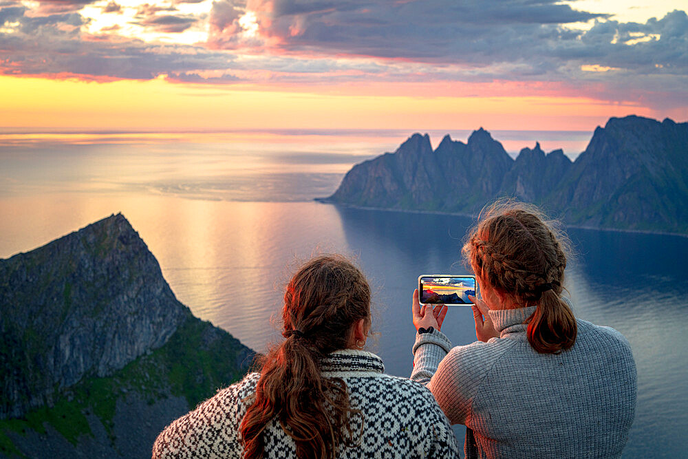 Two young Scandinavian women photographing the fjord at sunset with smartphone, Senja island, Troms county, Norway, Scandinavia, Europe