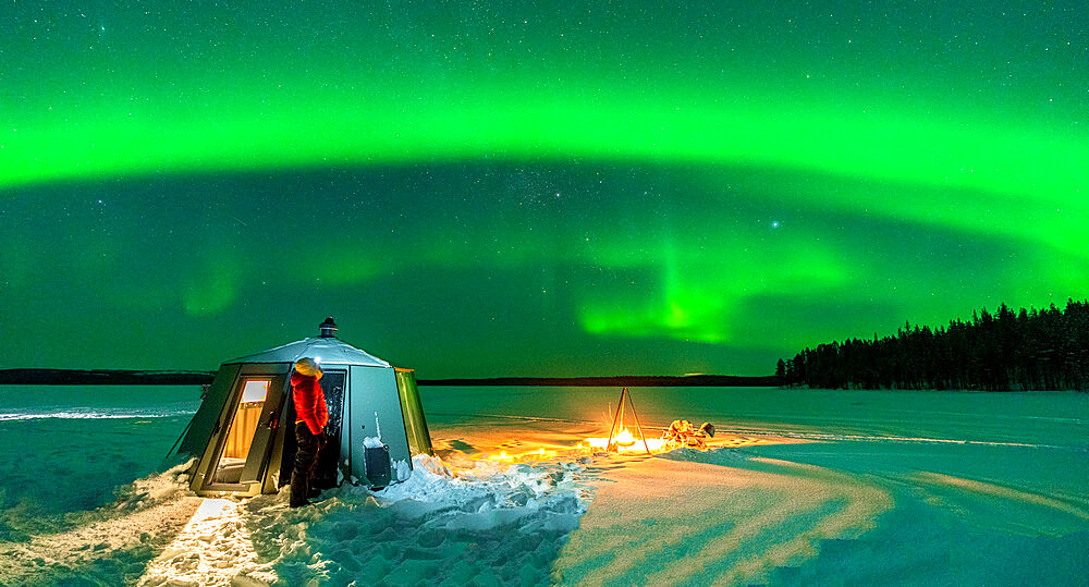 Hiker watching the Aurora Borealis (Northern Lights) close to bonfire and igloo in the frozen landscape, Jokkmokk, Lapland, Sweden, Scandinavia, Europe