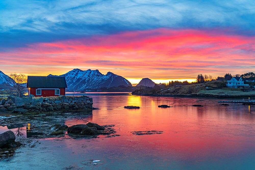 Fiery sky at sunrise over a red Rorbu facing the frozen sea, Ballstad, Vestvagoy, Lofoten Islands, Norway