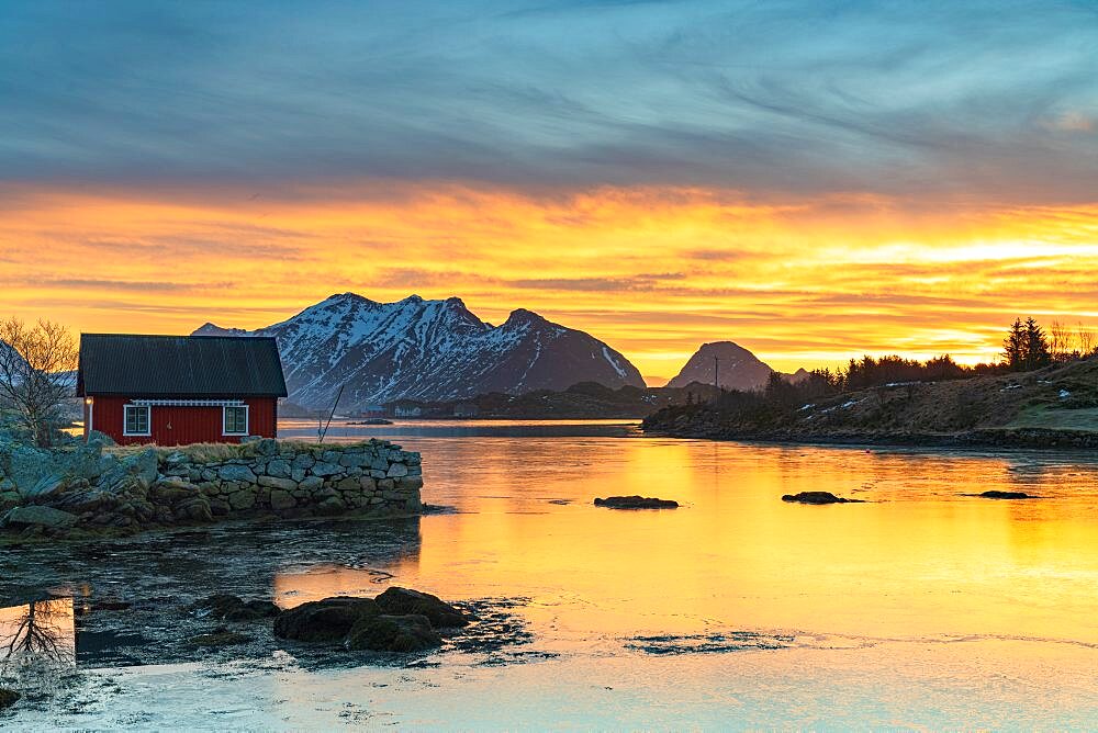 Snowcapped mountains and fisherman's red cabin under the burning sky at dawn, Ballstad, Vestvagoy, Lofoten Islands, Norway