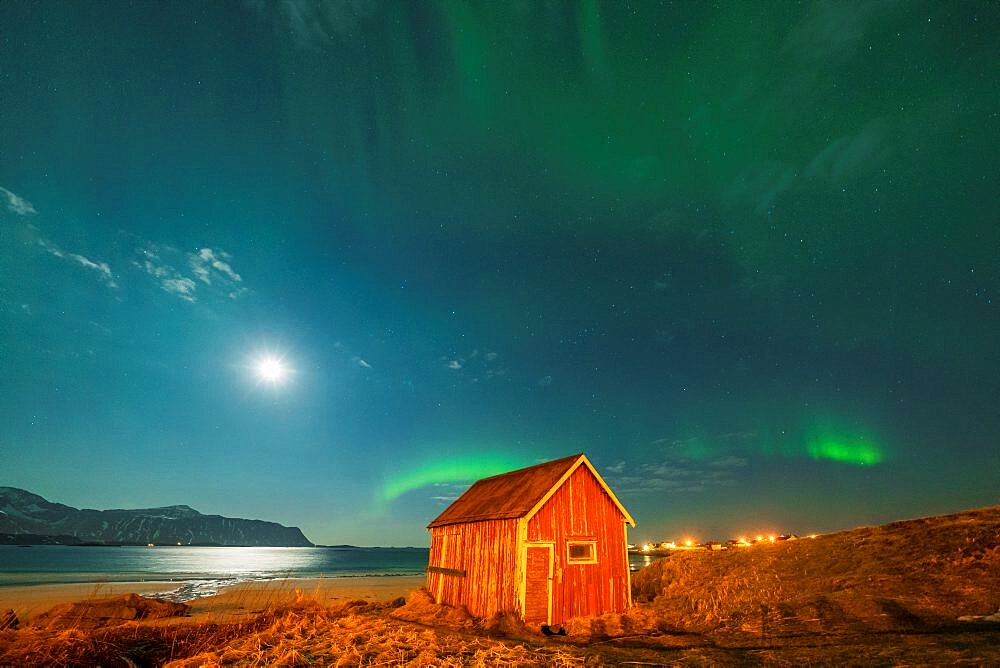 Red wood cabin on sand beach lit by moon during the Northern Lights, Ramberg, Nordland county, Lofoten Islands, Norway