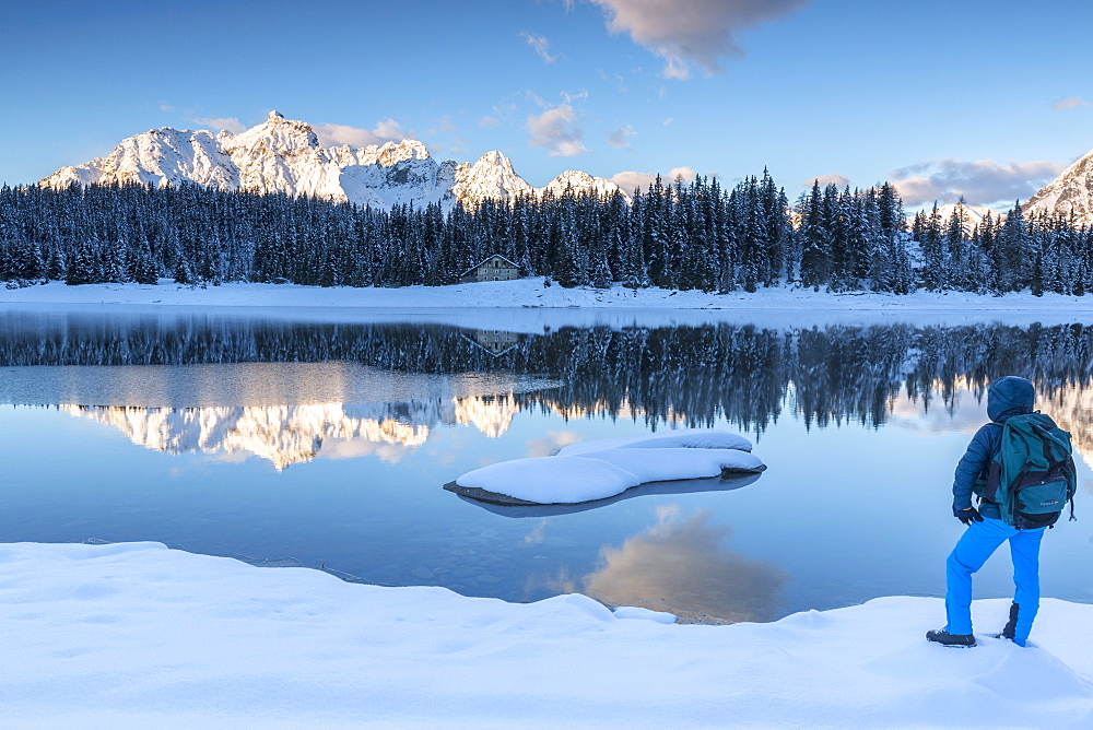 Hiker admires the snowy peaks and woods reflected in Lake Palu at dawn, Malenco Valley, Valtellina, Lombardy, Italy, Europe