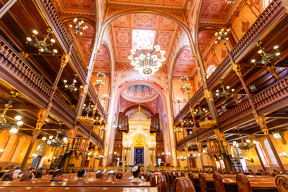 Wide-angle view inside the Dohány Street Synagogue, showing ornate chandeliers, wooden balconies, and the ark. Horizontal landscape orientation.