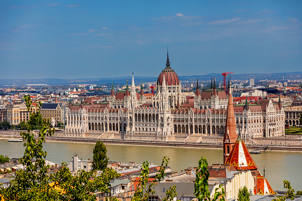 Elevated wide-angle view of the Hungarian Parliament Building on the Danube riverbank, seen from Buda with surrounding rooftops. Horizontal landscape orientation.