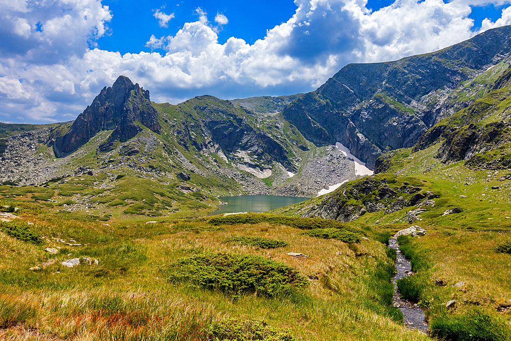 Horizontal, eye-level shot – Meadow and small stream in foreground with peaks and lake in background. Rila Mountains, Bulgaria.