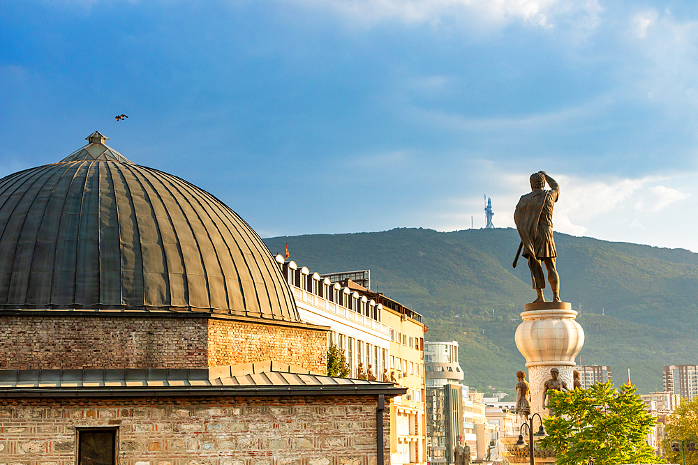 Horizontal eye-level shot of a domed stone building and a large statue of a man holding a sword, set against a backdrop of mountains and a cloudy blue sky in Skopje, North Macedonia.